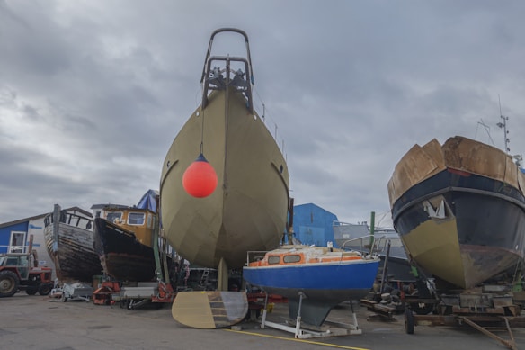 A shipyard scene with several boats on land, including a large boat with a bright red buoy hanging from it in the center. On the left, there is an older wooden boat, and on the right, a black and brown vessel. A small blue sailboat is in front, and various equipment and a red tractor can be seen in the foreground, with a cloudy sky above.