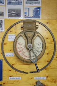 A vintage engine order telegraph mounted on a wooden board, displaying various power settings in both red and white segments. The telegraph has a metal handle and is surrounded by several black-and-white photographs of ships. Labels are attached nearby, providing additional information in a foreign language.