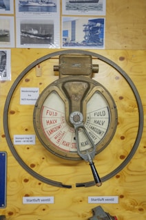 A vintage engine order telegraph mounted on a wooden board, displaying various power settings in both red and white segments. The telegraph has a metal handle and is surrounded by several black-and-white photographs of ships. Labels are attached nearby, providing additional information in a foreign language.