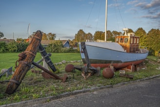 A boat is displayed on dry land, surrounded by various maritime artifacts, including large rusty anchors and buoy balls. The boat appears to be old and weathered, with a wooden cabin and a white and blue hull. There is a grassy area in the background, along with some houses and trees under a clear sky.