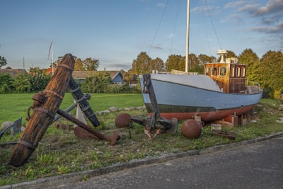 A boat is displayed on dry land, surrounded by various maritime artifacts, including large rusty anchors and buoy balls. The boat appears to be old and weathered, with a wooden cabin and a white and blue hull. There is a grassy area in the background, along with some houses and trees under a clear sky.