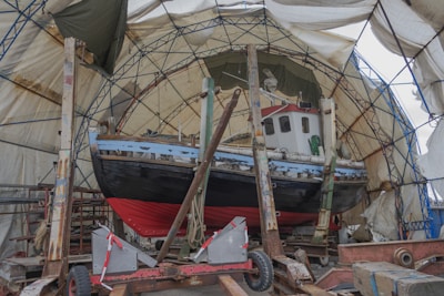 A boat is positioned inside a covered structure, likely for repair or maintenance. The boat has a red and black hull with a white cabin. It is supported by wooden and metal braces, and the covering structure consists of a metal frame with translucent fabric draped over it.