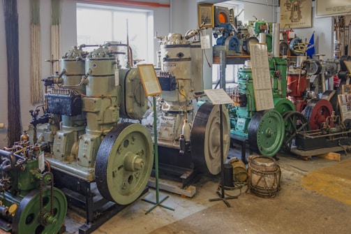 An indoor exhibit displays a collection of vintage industrial engines. The engines, varying in size and design, are lined up side by side, each mounted on platforms. Plaques with information are placed on stands in front of some of the engines. The room is well-lit with natural light coming through a window, and various components and machinery parts are visible in the background.