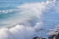 Powerful ocean waves crash against the shore, creating a dynamic spray of water. The coastline is lined with rocks which the waves are breaking over, and the ocean stretches into the distance with its vibrant blue color.
