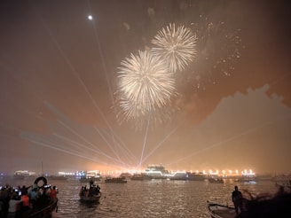 Fireworks light up the night sky over a large body of water, with boats and people watching the display. Bright lights and laser beams enhance the festive atmosphere.