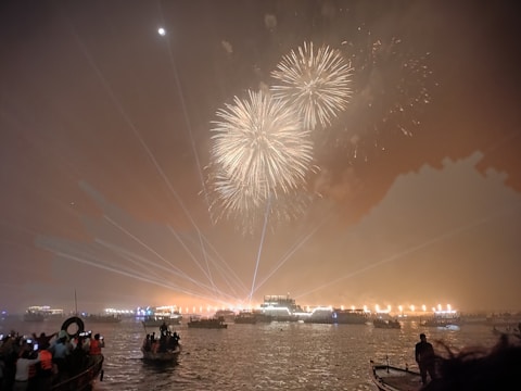 Fireworks light up the night sky over a large body of water, with boats and people watching the display. Bright lights and laser beams enhance the festive atmosphere.
