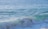 A sunlit ocean with gentle, rolling waves. The water is a beautiful blend of blue and turquoise shades, with white foam forming on the wave crests.