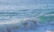 A sunlit ocean with gentle, rolling waves. The water is a beautiful blend of blue and turquoise shades, with white foam forming on the wave crests.