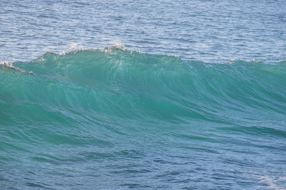 A clear ocean wave with a vibrant turquoise color, cresting with slight foam on its top. The sea appears calm in the background with varying shades of blue.