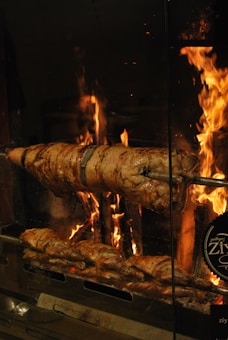 A large piece of meat is being roasted over an open flame. The meat is skewered on a rotisserie, which allows it to cook evenly as it turns slowly above the fire. Flames and glowing embers create a warm and rustic ambiance, emphasizing the cooking process.