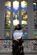 A sculpture stands in the foreground with intricate, abstract features. Behind it, vibrant stained glass windows showcase nature scenes with lush greenery, a waterfall, and bright skies. White string lights form a delicate backdrop across the lower half of the image.