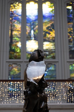 A sculpture stands in the foreground with intricate, abstract features. Behind it, vibrant stained glass windows showcase nature scenes with lush greenery, a waterfall, and bright skies. White string lights form a delicate backdrop across the lower half of the image.