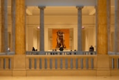 A museum interior with classical architecture featuring large stone columns and a sculpture of three figures in the background. Several people are observing the art piece, some standing while others appear seated. The lighting is warm and creates a serene atmosphere.