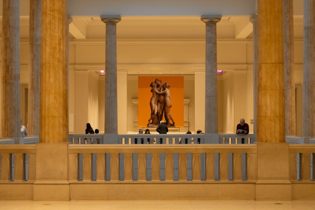 A museum interior with classical architecture featuring large stone columns and a sculpture of three figures in the background. Several people are observing the art piece, some standing while others appear seated. The lighting is warm and creates a serene atmosphere.