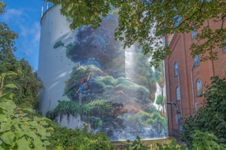 A large cylindrical structure adorned with a vibrant mural depicting a child with a backpack reaching toward a lush, green landscape with birds flying overhead. The cylinder is situated next to a red brick building, partially obscured by trees and foliage.