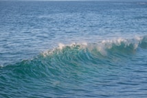 A clear ocean wave is cresting in the foreground, with sunlight reflecting off its surface, creating a mix of blue and green hues. The ocean extends into the distance, showcasing a calm and expansive water surface.