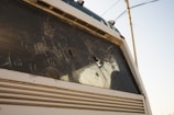 A close-up view of a vehicle with a shattered windshield, showing multiple cracks and holes. The vehicle appears to be stationary, with visible damage and possibly no occupants inside. The background includes a utility pole and wires against a pale blue sky.