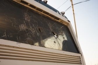 A close-up view of a vehicle with a shattered windshield, showing multiple cracks and holes. The vehicle appears to be stationary, with visible damage and possibly no occupants inside. The background includes a utility pole and wires against a pale blue sky.