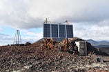 A remote, rocky terrain with solar panels mounted on a metal frame. The ground is covered with red and brown volcanic rocks. In the background, there are rugged hills and a cloudy sky.