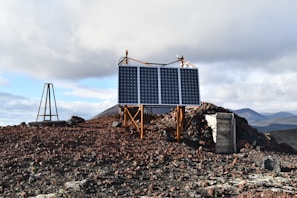 A remote, rocky terrain with solar panels mounted on a metal frame. The ground is covered with red and brown volcanic rocks. In the background, there are rugged hills and a cloudy sky.