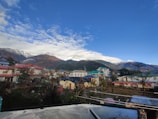 A small mountain town with colorful buildings in the foreground. Snow-capped mountains and a vibrant blue sky with some clouds can be seen in the background. Several residential houses, trees, and electrical lines are visible, surrounded by a serene and scenic environment.
