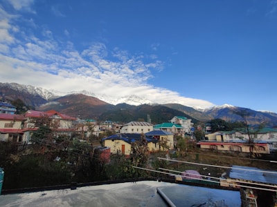 A small mountain town with colorful buildings in the foreground. Snow-capped mountains and a vibrant blue sky with some clouds can be seen in the background. Several residential houses, trees, and electrical lines are visible, surrounded by a serene and scenic environment.