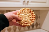 A hand holds a waffle featuring a smiling gingerbread man design. The waffle is a light golden brown, with a detailed imprint of the gingerbread man including facial features and buttons. The background shows a kitchen setting with white cabinets and a tiled backsplash.