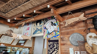 A wooden ceiling adorned with various artistic elements, including painted panels featuring traditional Japanese art and masks. Two carved wooden fish hang from above, while other decorative items, such as woven and wooden accents, are arranged on the walls and surfaces.