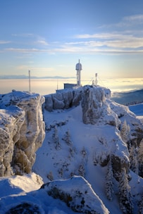A snow-covered mountainous landscape with a weather station or observatory perched on top of a rugged cliff. The structure features antennas and a dome, likely for scientific observation. Pine trees dusted with snow line the rocky outcrop, and a vast expanse of clouds stretches across the horizon under a clear sky.