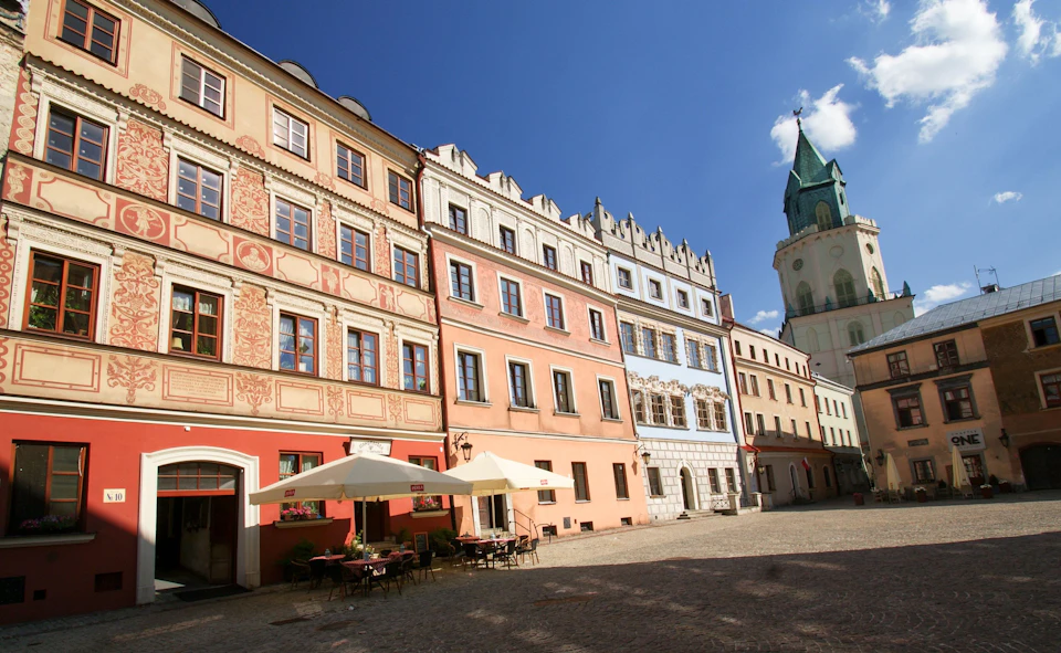 The image depicts a charming cobblestone square in a European town, with buildings painted in various colors and architectural styles lining the street. The square is filled with people walking around, adding to its lively atmosphere. In the background, there's a church with a green spire that stands out against the clear blue sky dotted with white clouds.

The square features several outdoor seating areas, including tables and chairs under umbrellas, where visitors can enjoy their meals or relax in the pleasant weather. The buildings showcase a range of architectural styles, from traditional to more modern designs, creating an interesting visual contrast within the scene.