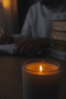 A lit candle in the foreground creates a warm, intimate glow. In the background, a person is writing in a notebook, surrounded by a stack of books. The lighting is dim, enhancing the cozy atmosphere.