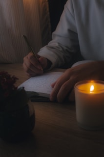 A person is writing in a notebook with a pen on a wooden table. A lit candle provides soft illumination, creating a warm and cozy atmosphere. There is also a plant placed on the table, adding a touch of greenery.
