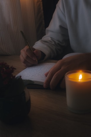 A person is writing in a notebook with a pen on a wooden table. A lit candle provides soft illumination, creating a warm and cozy atmosphere. There is also a plant placed on the table, adding a touch of greenery.