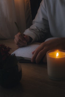 A person is writing in a notebook with a pen on a wooden table. A lit candle provides soft illumination, creating a warm and cozy atmosphere. There is also a plant placed on the table, adding a touch of greenery.
