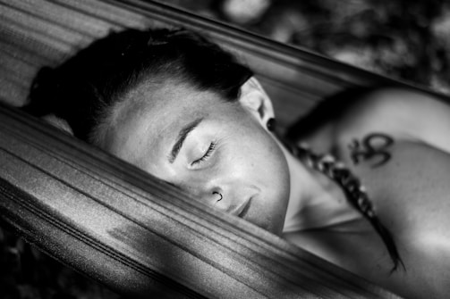 A person with braided hair and a small nose ring rests peacefully in a hammock. There's a visible tattoo on the shoulder featuring the Om symbol. The image is in black and white, highlighting the serene and relaxed expression on their face.