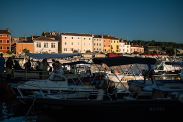 A picturesque coastal scene with colorful buildings lining the waterfront, casting reflections on the water. Boats with canopies are docked in the harbor, and people are gathered on the boats and the dock, enjoying the view. The sky is clear, adding to the vibrant atmosphere.