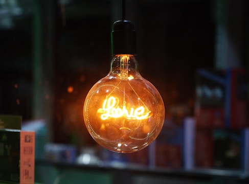 A glowing lightbulb with the word 'Love' written inside, illuminating its surroundings with a warm amber glow, set against a dark, blurred background.