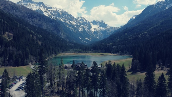 Nestled between towering snow-capped mountains, a serene lake reflects the lush green forests that surround it. A small cluster of buildings sits near the edge of the water, and a line of vehicles is parked along a winding road in the foreground. The sky is partly cloudy, accentuating the tranquil atmosphere of the landscape.