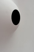 A minimalistic design with a large white semi-spherical dome featuring a single, featureless black oval shape in the center. The background is a plain, light grey color making the black shape stand out starkly against the otherwise neutral tones.