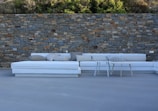 A spacious outdoor seating area with a white L-shaped sofa equipped with various pillows, positioned in front of a rustic stone wall. Two small coffee tables are placed in front of the sofa. The ground is covered in a smooth gray flooring material with a backdrop of greenery above the stone wall.