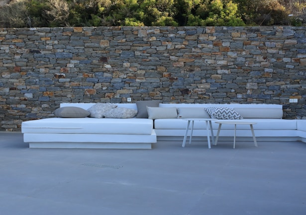 A spacious outdoor seating area with a white L-shaped sofa equipped with various pillows, positioned in front of a rustic stone wall. Two small coffee tables are placed in front of the sofa. The ground is covered in a smooth gray flooring material with a backdrop of greenery above the stone wall.