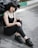 A young person is sitting on the ground next to a motorcycle, wearing all black attire including a tank top, jeans, and sneakers. They have a distinctive gothic style with dark makeup and layered silver necklaces.