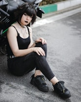 A young person is sitting on the ground next to a motorcycle, wearing all black attire including a tank top, jeans, and sneakers. They have a distinctive gothic style with dark makeup and layered silver necklaces.
