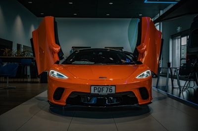 A sleek, bright orange sports car is displayed in a showroom with its doors open upward. The car sits on a tiled floor under soft indoor lighting, and there are plants and furniture visible in the background.