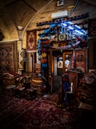 A beautifully decorated interior with an array of colorful, intricately patterned rugs hanging on the walls and covering the floor. The dim lighting creates a warm and cozy atmosphere, with the soft glow highlighting the textures and details of each rug. A doorway in the center reveals additional displays in a well-lit adjacent room.