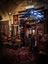 A beautifully decorated interior with an array of colorful, intricately patterned rugs hanging on the walls and covering the floor. The dim lighting creates a warm and cozy atmosphere, with the soft glow highlighting the textures and details of each rug. A doorway in the center reveals additional displays in a well-lit adjacent room.