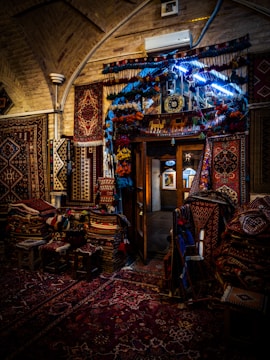A beautifully decorated interior with an array of colorful, intricately patterned rugs hanging on the walls and covering the floor. The dim lighting creates a warm and cozy atmosphere, with the soft glow highlighting the textures and details of each rug. A doorway in the center reveals additional displays in a well-lit adjacent room.