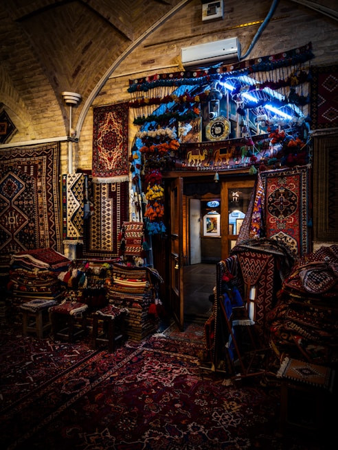 A beautifully decorated interior with an array of colorful, intricately patterned rugs hanging on the walls and covering the floor. The dim lighting creates a warm and cozy atmosphere, with the soft glow highlighting the textures and details of each rug. A doorway in the center reveals additional displays in a well-lit adjacent room.