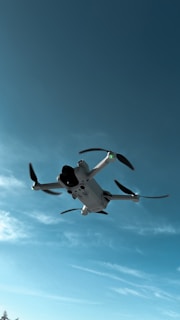 A drone flies upward against a blue sky, with its rotor blades spinning and casting small shadows. The drone appears to be compact with a sleek design, featuring a camera on its front. The backdrop includes a few wispy clouds.