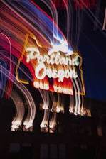 A neon sign with glowing white and red lettering spells out 'Portland, Oregon' and 'Old Town'. Above the text, a white outline of a deer is lit up. The image features motion blur, creating streaks of light across the dark night background, adding a dynamic and vibrant effect.